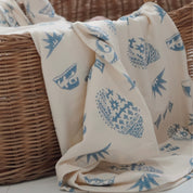 Cream and blue Kailani swaddle featuring beach-inspired shell and wave print, draped over a wicker basket.