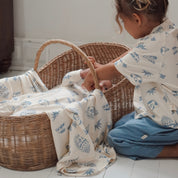 Older sibling gently interacting with baby in a basket wrapped in the Kailani swaddle, both wearing matching prints.
