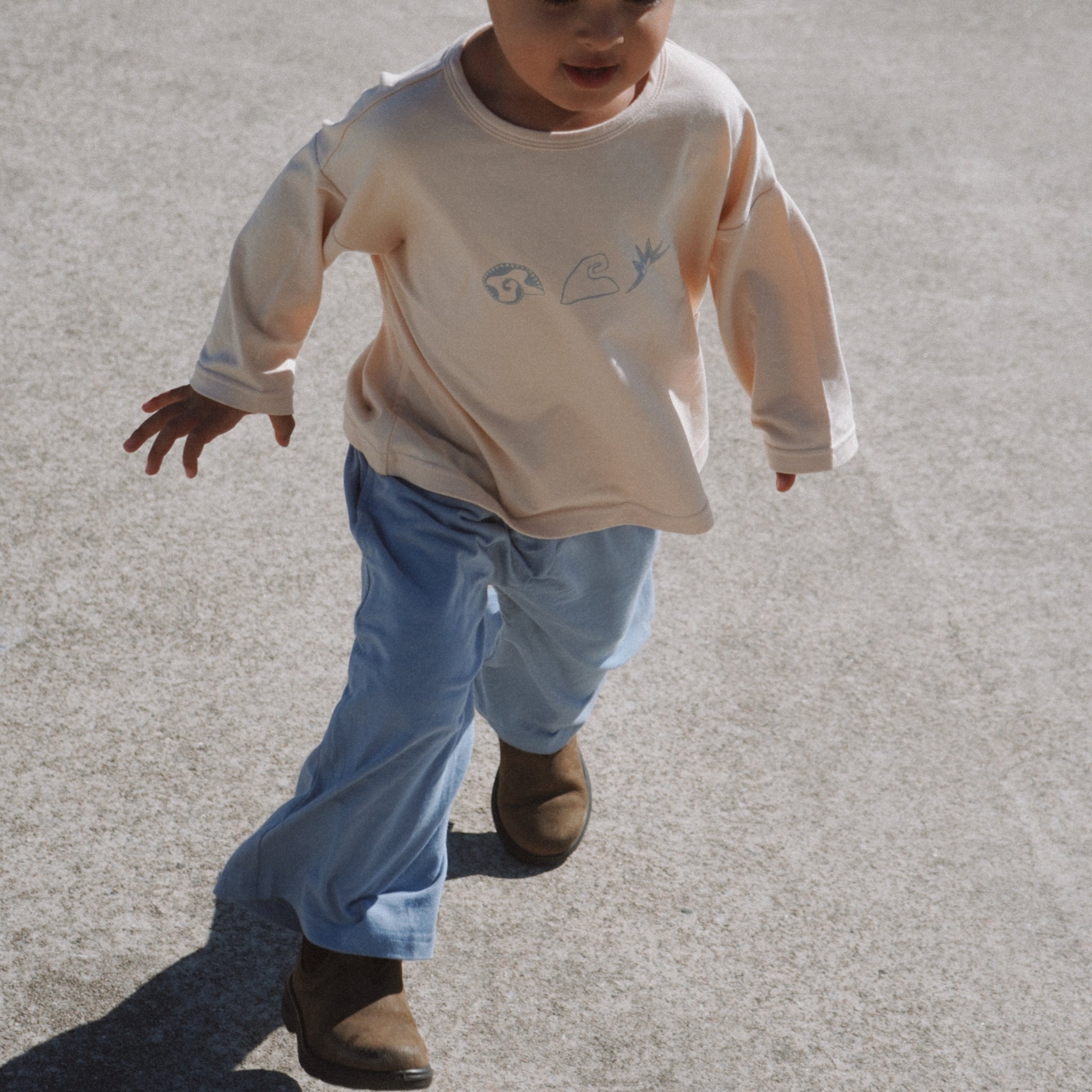 Child wearing a beige long-sleeve shirt and blue pants walking on a concrete surface.