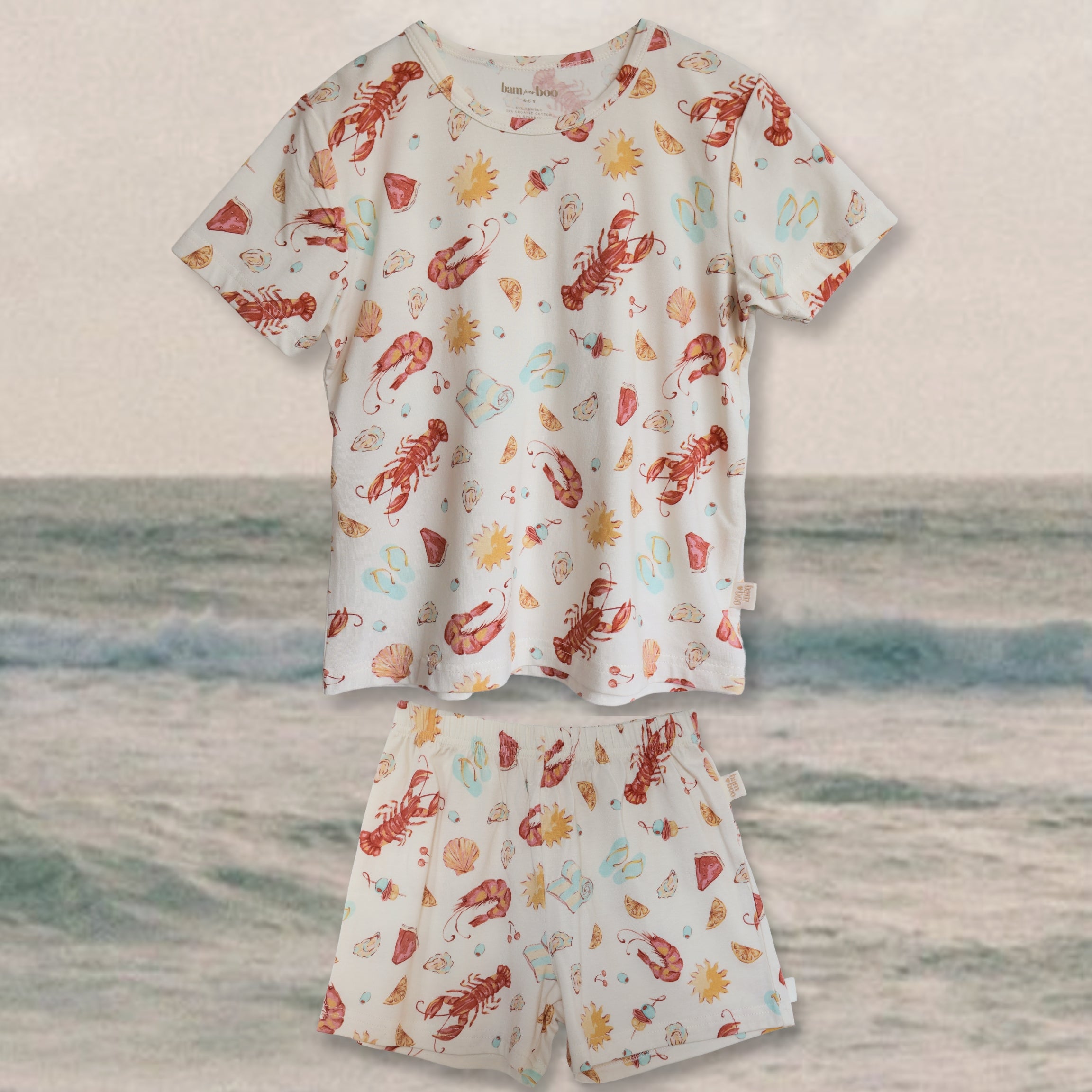 kids Pajamas in cream with Aussie summer print (lobsters, prawns, shells, sun, thongs); long sleeves; beach backdrop.
