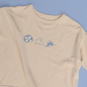 Flat lay of Beachcomber tee on soft blue background, highlighting front shell, wave, and sun illustrations.