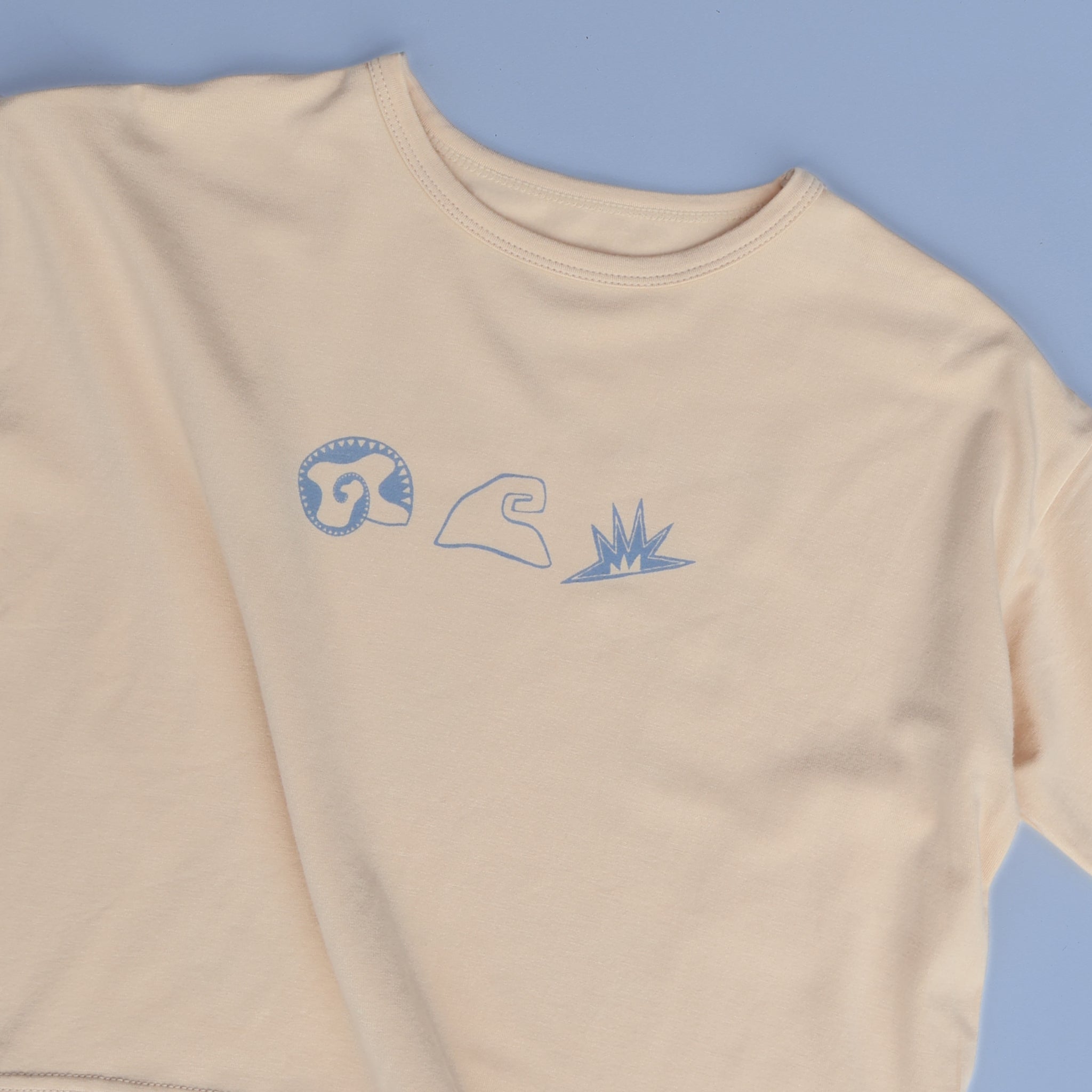 Flat lay of Beachcomber tee on soft blue background, highlighting front shell, wave, and sun illustrations.