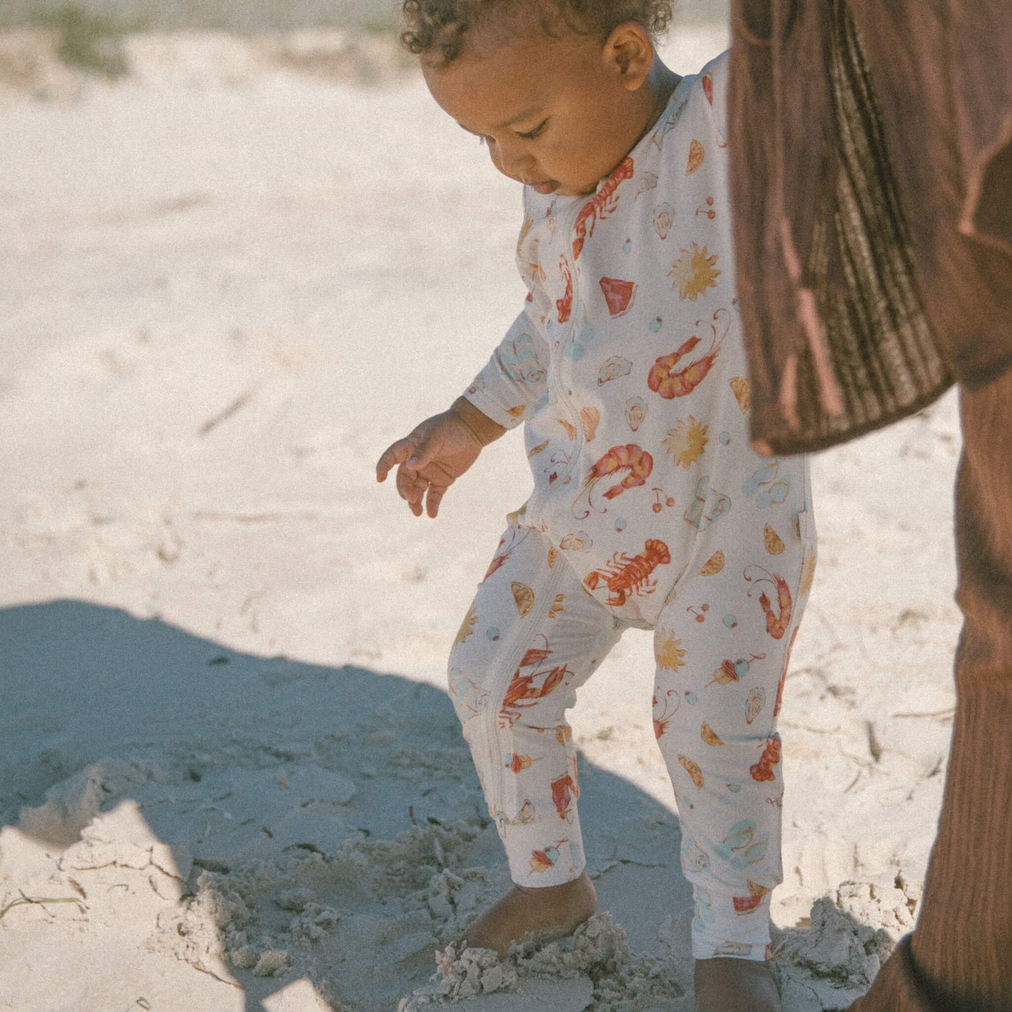 Baby wearing cream Australian-summer printed bamboo onesie, playing on sand outdoors.