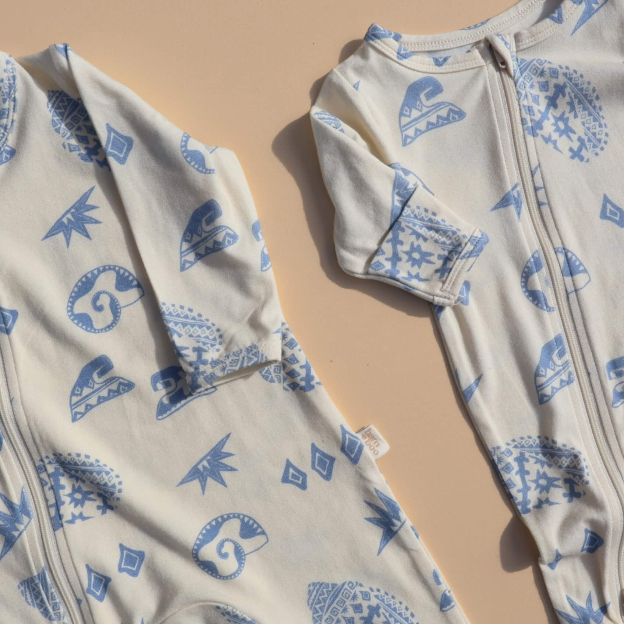 Kailani baby zip suit and matching swaddle displayed flat on neutral peach surface, showcasing blue coastal illustrations.