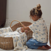 Toddler adjusting a swaddle over baby in basket, both dressed in Kailani print with cream and blue coastal motifs.