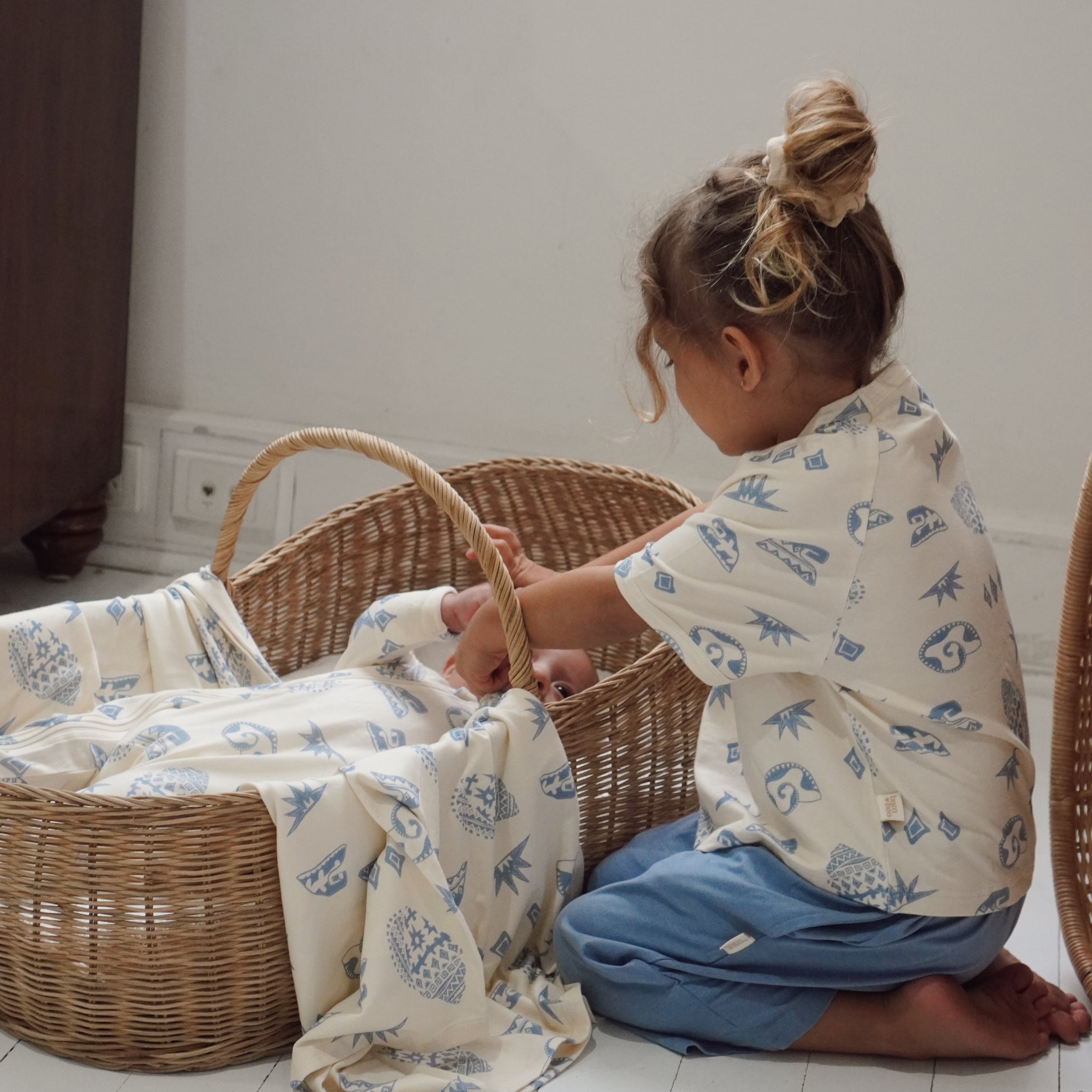 Toddler adjusting a swaddle over baby in basket, both dressed in Kailani print with cream and blue coastal motifs.