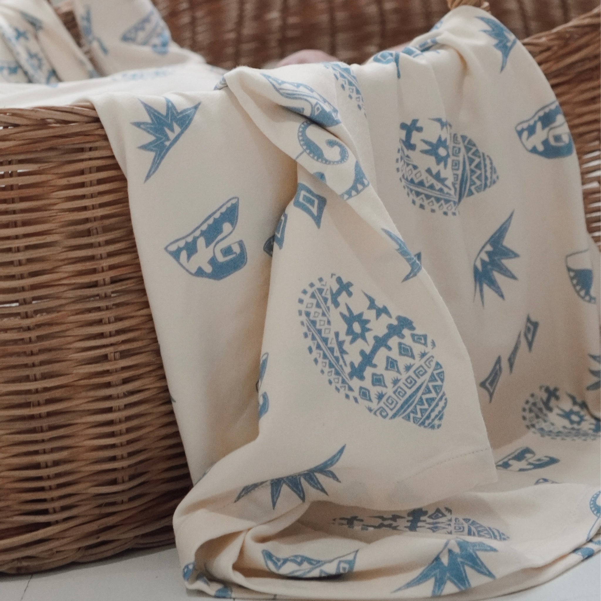 Cream and blue Kailani swaddle featuring beach-inspired shell and wave print, draped over a wicker basket.