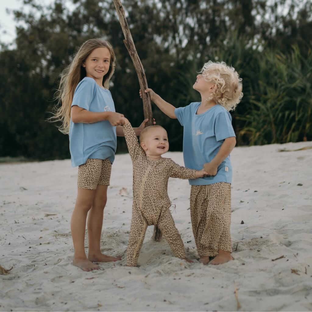 Three children in matching blue shirts and leopard print pants standing on a sandy beach.