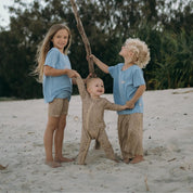 Three children in matching blue shirts and leopard print pants standing on a sandy beach.