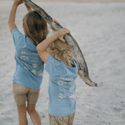 Two children in matching blue shirts with beach-themed designs on a sandy beach.