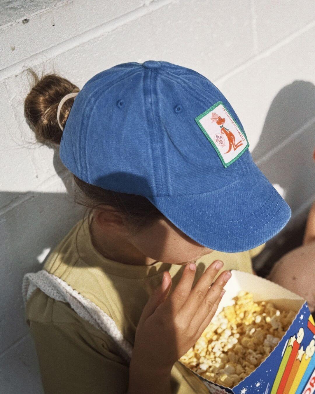 Person wearing a blue cap with a logo, eating popcorn from a box.