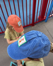Blue cap with a patch worn by a child, with another child wearing an orange cap in the background.