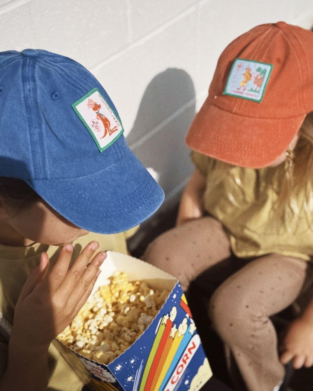 Two children wearing baseball caps with patches, sitting next to a box of popcorn.