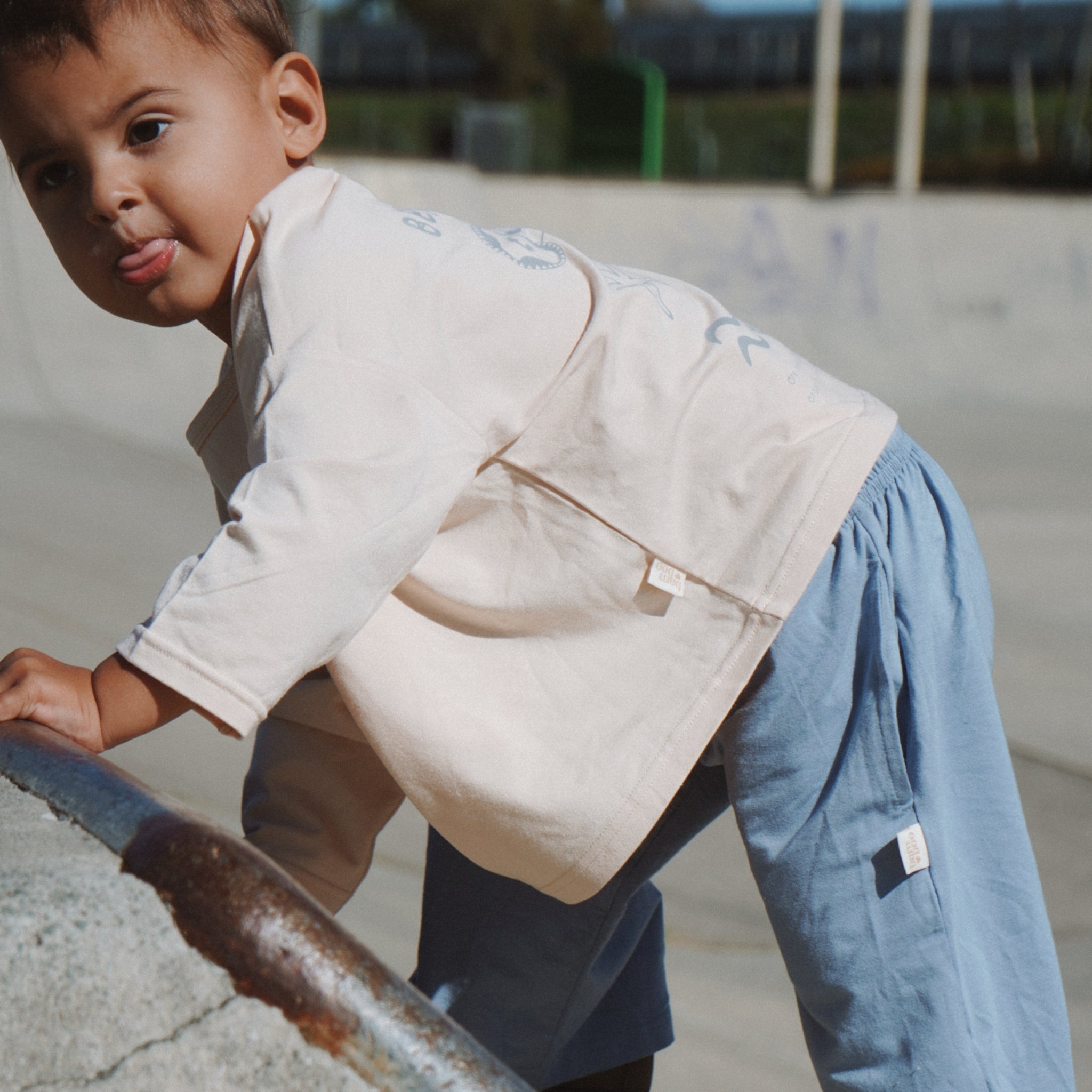 Child in a beige vest and blue pants climbing on a metal railing outdoors.