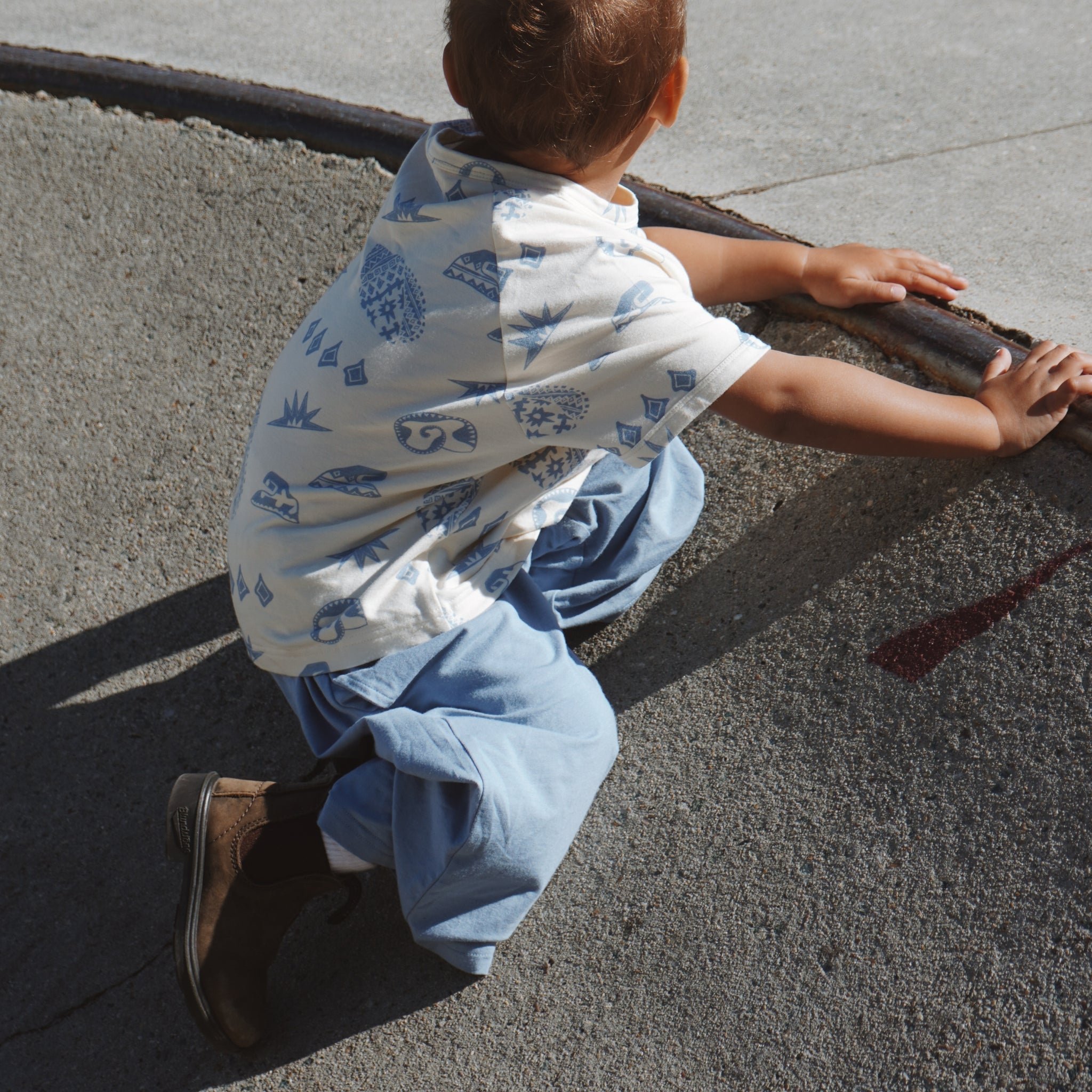 Child wearing Kailani print tee with soft blue shells and waves, crouched on pavement in natural sunlight.