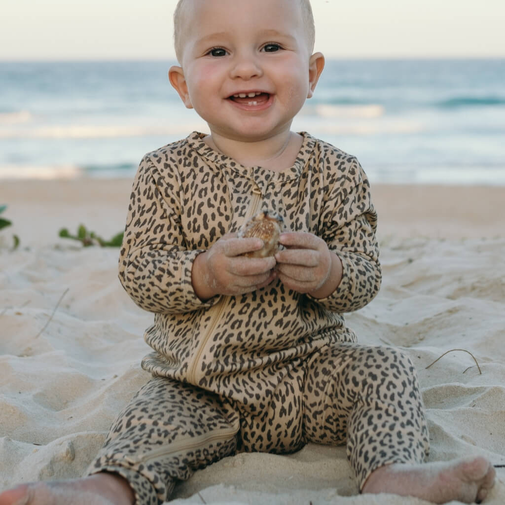 Baby in leopard print jumpsuit sitting on a sandy beach.
