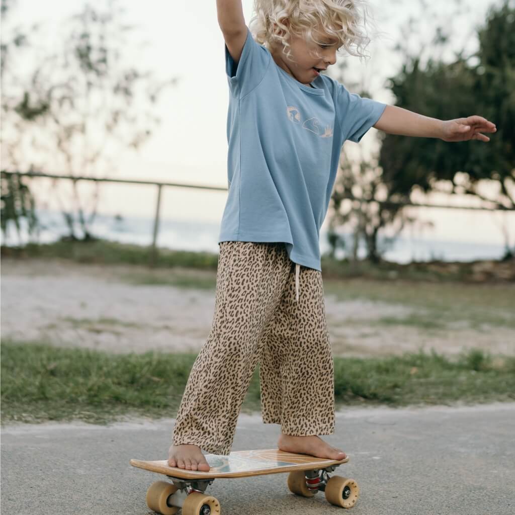 Child in a blue shirt and leopard print pants standing on a skateboard outdoors.