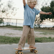 Child in a blue shirt and leopard print pants standing on a skateboard outdoors.