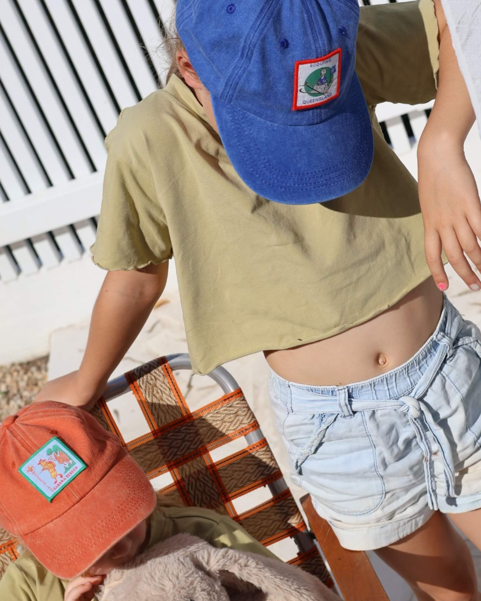 Two kids wearing blue and orange baseball caps with patches, sitting on a chair outdoors.