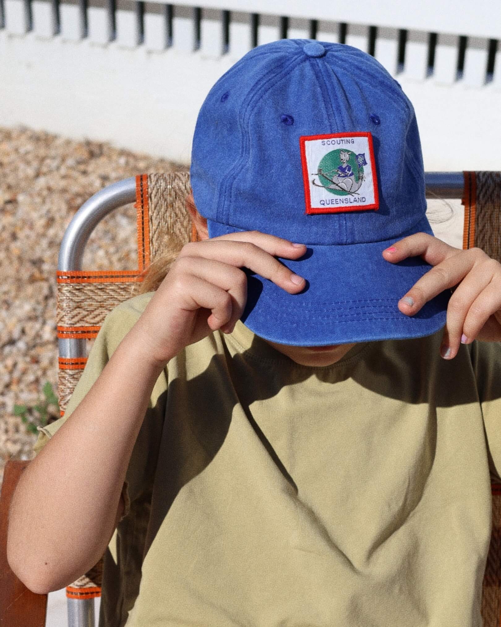 child wearing a blue cap with a Queensland patch, sitting outdoors.