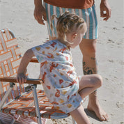 child wearing cream with Aussie summer print lobsters, prawns, shells, sun sitting on beach chair