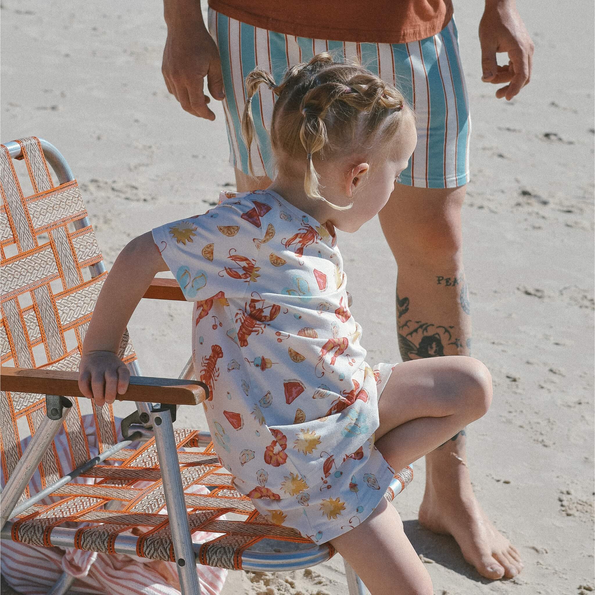 child wearing cream with Aussie summer print lobsters, prawns, shells, sun sitting on beach chair