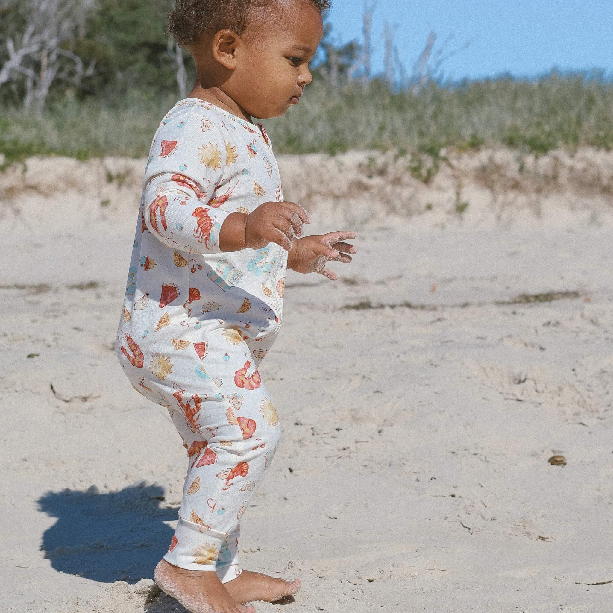 Side view of baby in long-sleeve romper with watercolor seaside icons, sandy beach background.
