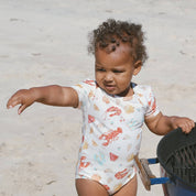 Short-sleeve baby onesie with watercolor seafood and beach icons on off-white, modeled at the beach.