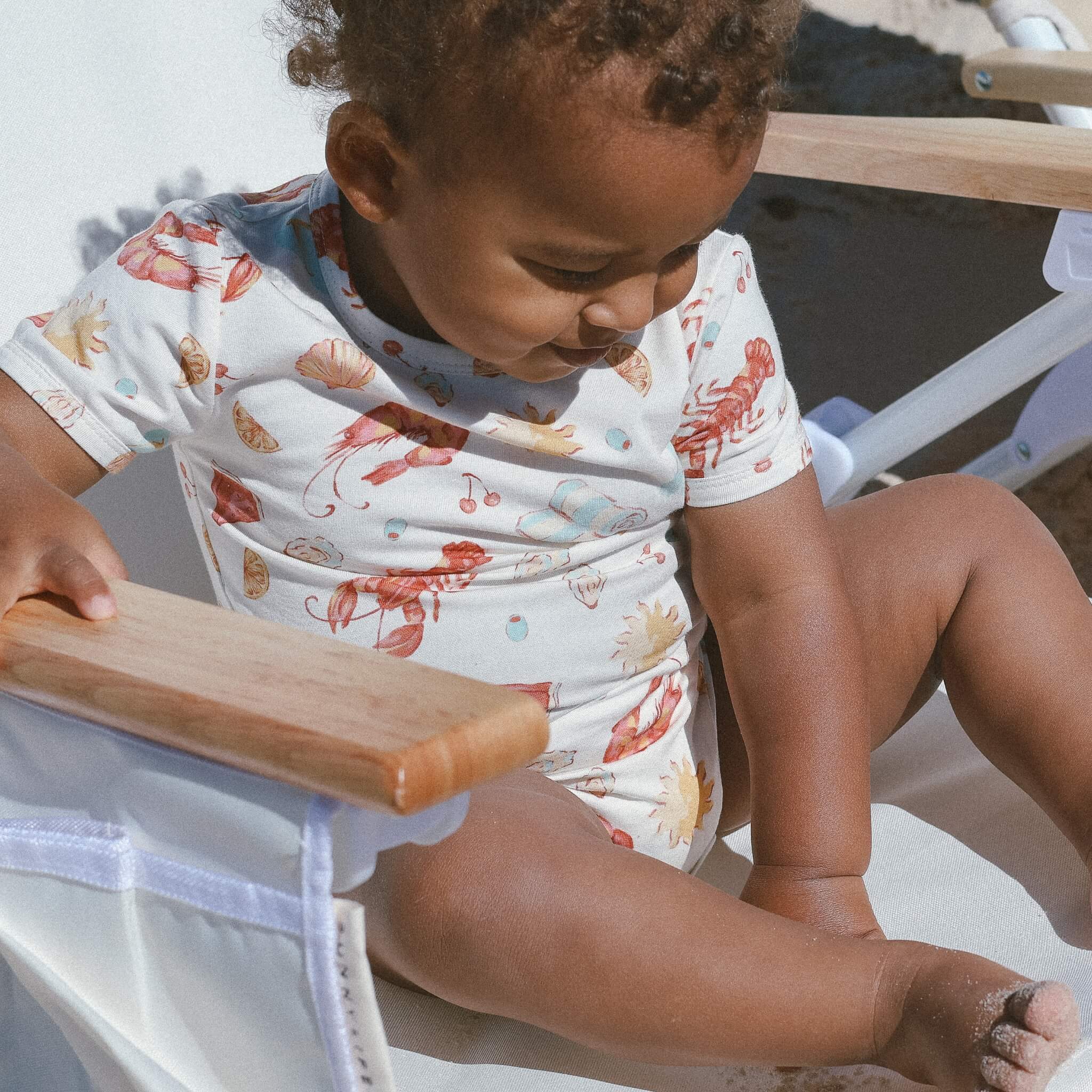 Close-up of toddler in cream Australian-christmas motif bodysuit; shoreline in background.