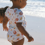 Infant bodysuit featuring playful seaside pattern—lobsters, prawns, shells, sun—cream base, ocean backdrop