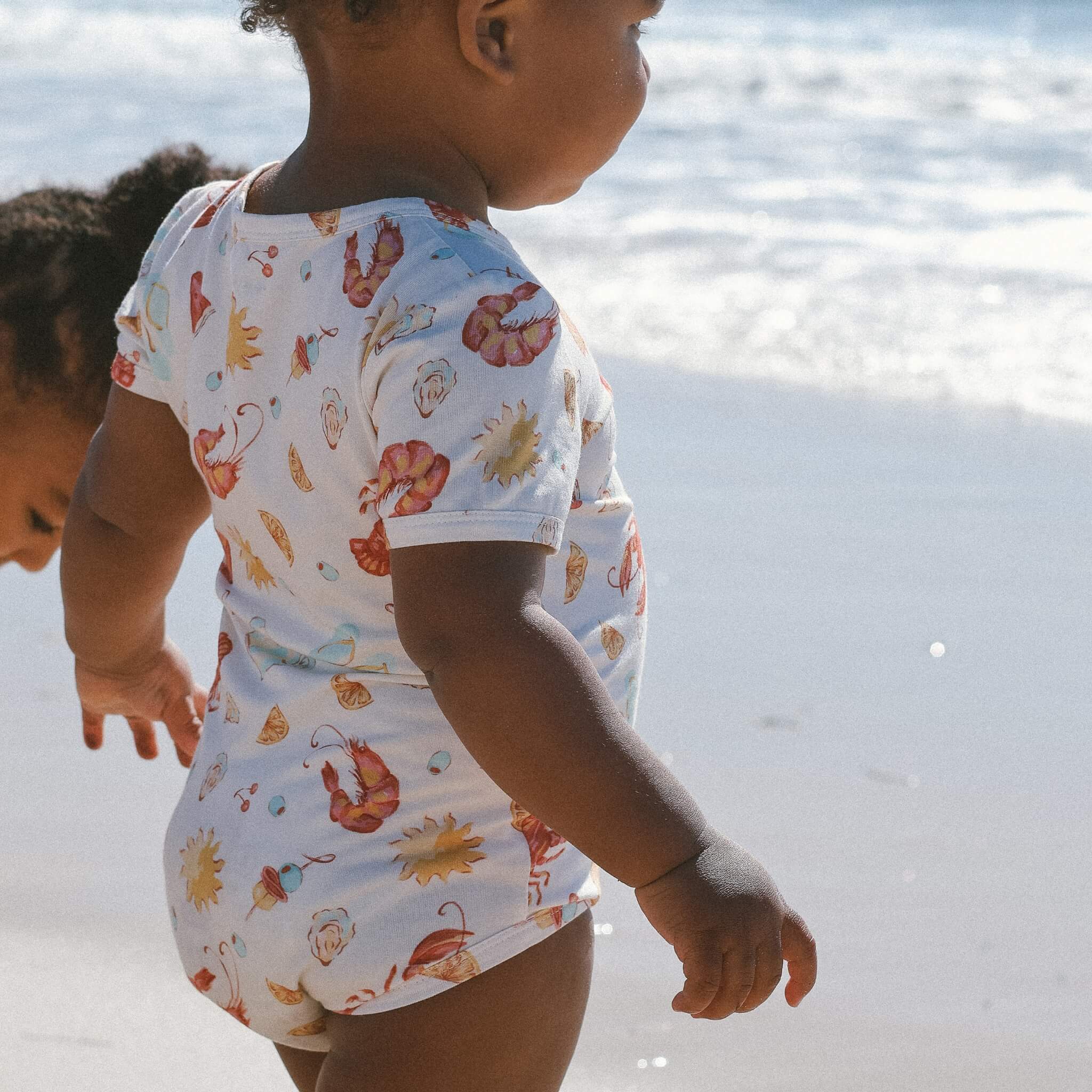 Infant bodysuit featuring playful seaside pattern—lobsters, prawns, shells, sun—cream base, ocean backdrop