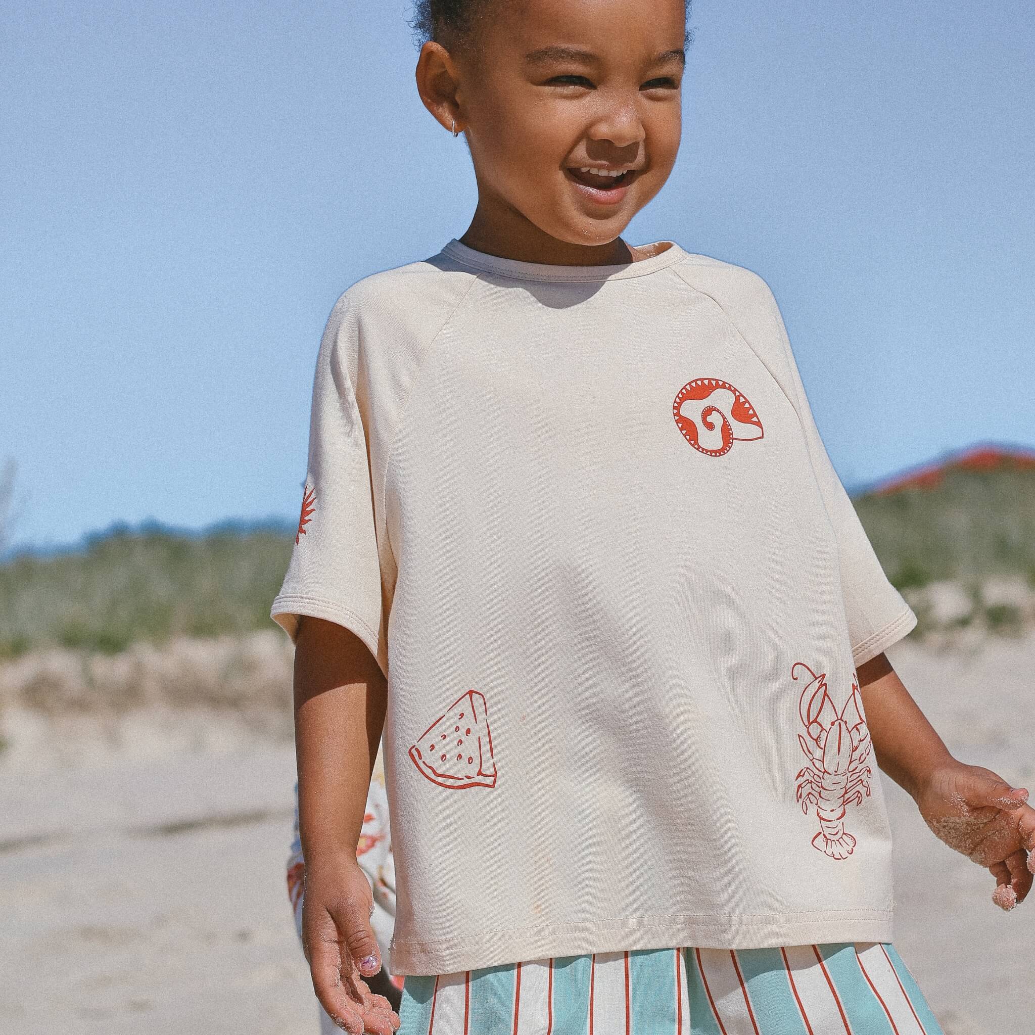 Child at the beach wearing cream oversized tee with red line-art motifs (shell, watermelon, lobster, sun)
