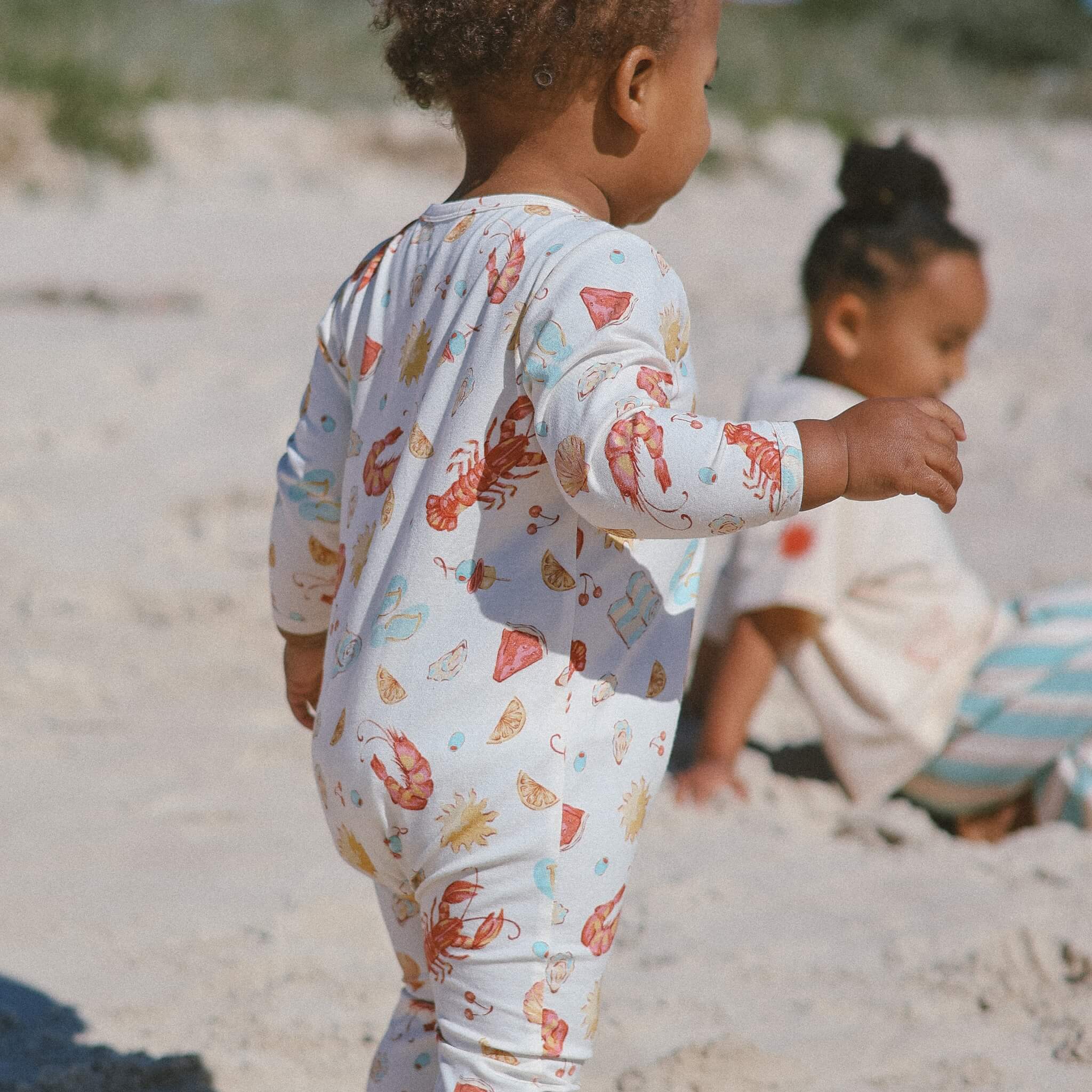 Infant wearing cream Australian-summer print onesie, playing on sand outdoors.