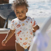 Baby wearing cream printed onesie “Aussie Christmas” pattern, beach setting.