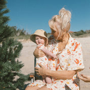 Matching family christmas outfits—cream seaside pattern—adult seated holding baby near a Christmas tree at the beach.