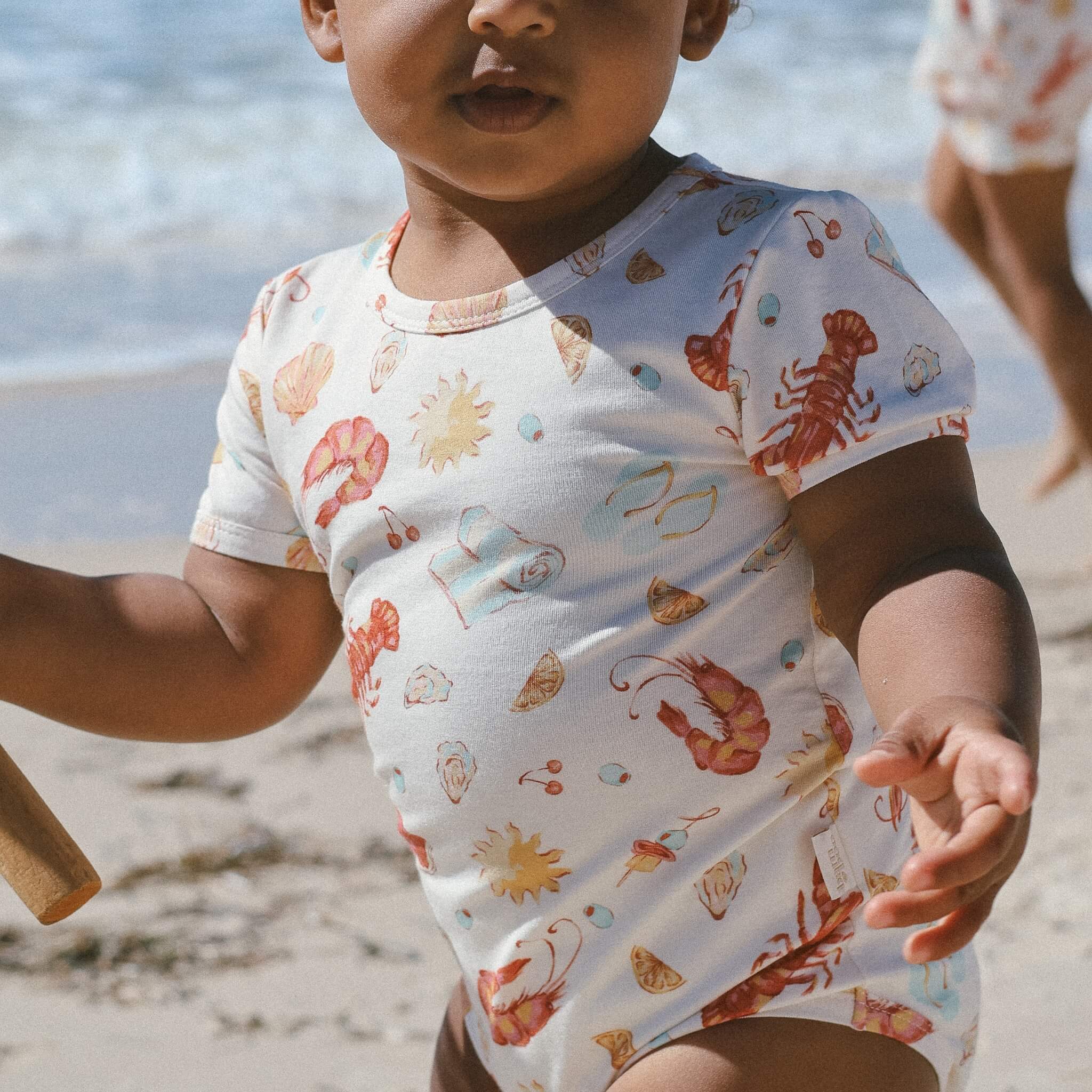 Baby wearing short-sleeve onesie in cream with Aussie summer print (lobsters, prawns, shells, sun) at the beach.