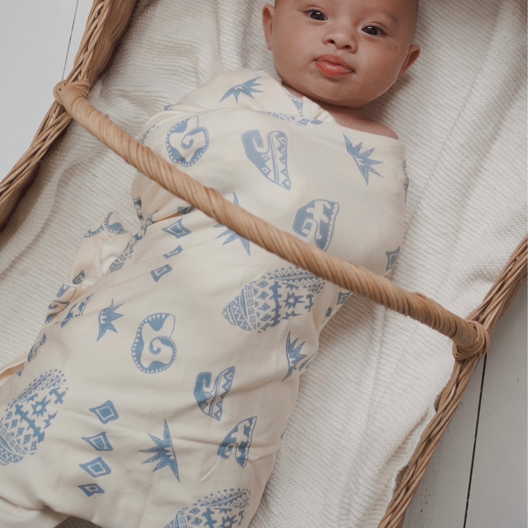 Newborn baby wrapped snugly in the Kailani swaddle, resting in a wicker bassinet.