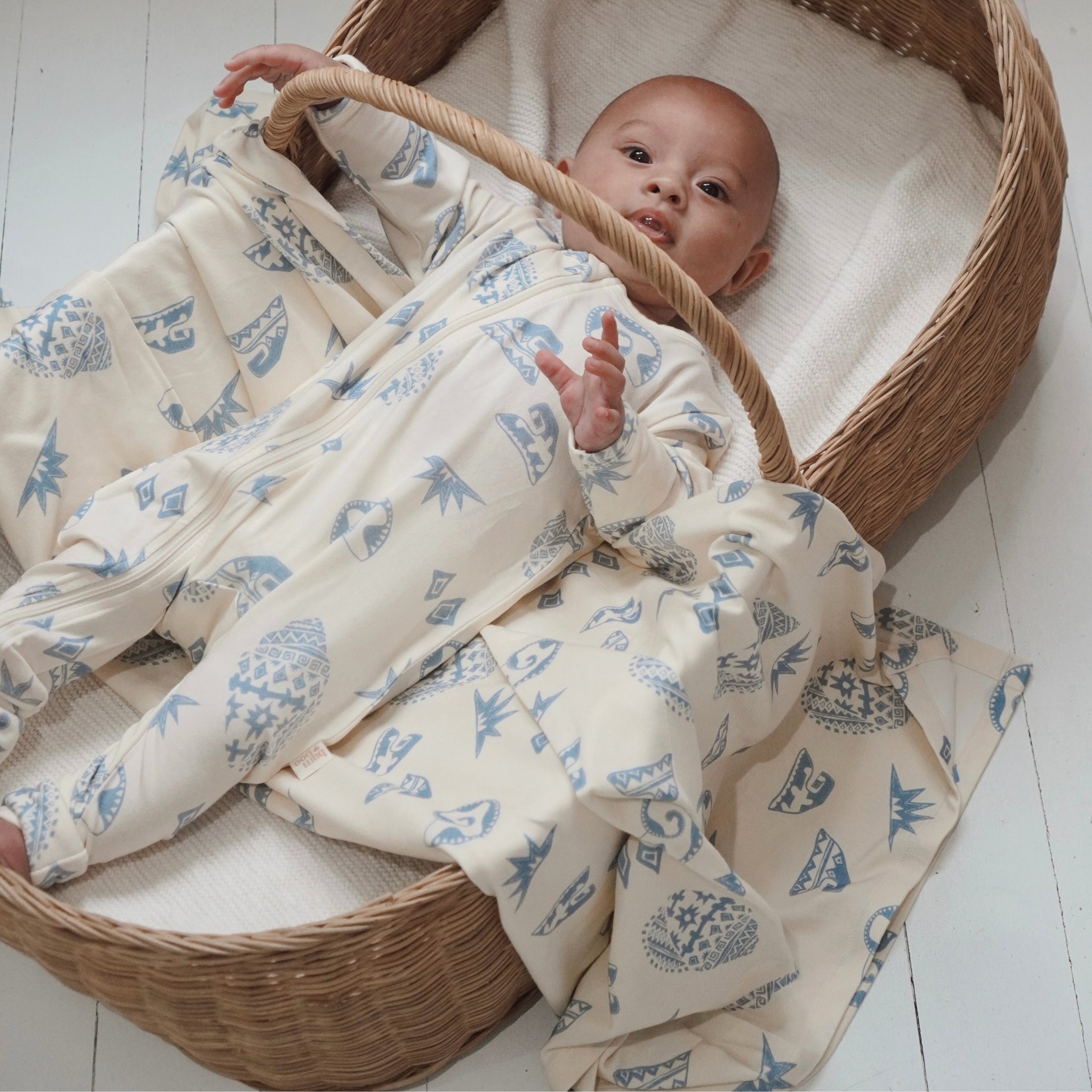 Baby in a wicker bassinet wearing Kailani zip suit, wrapped in matching coastal-print swaddle in cream and soft blue.