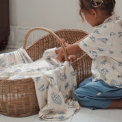 Older sibling gently interacting with baby in a basket wrapped in the Kailani swaddle, both wearing matching prints.