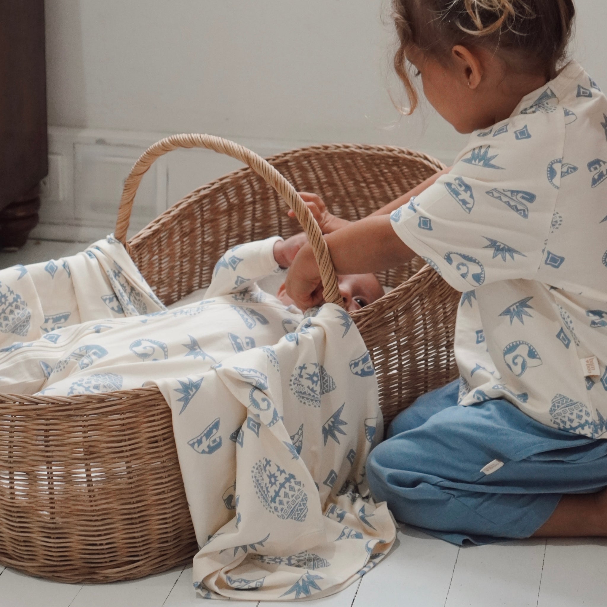 Older sibling gently interacting with baby in a basket wrapped in the Kailani swaddle, both wearing matching prints.