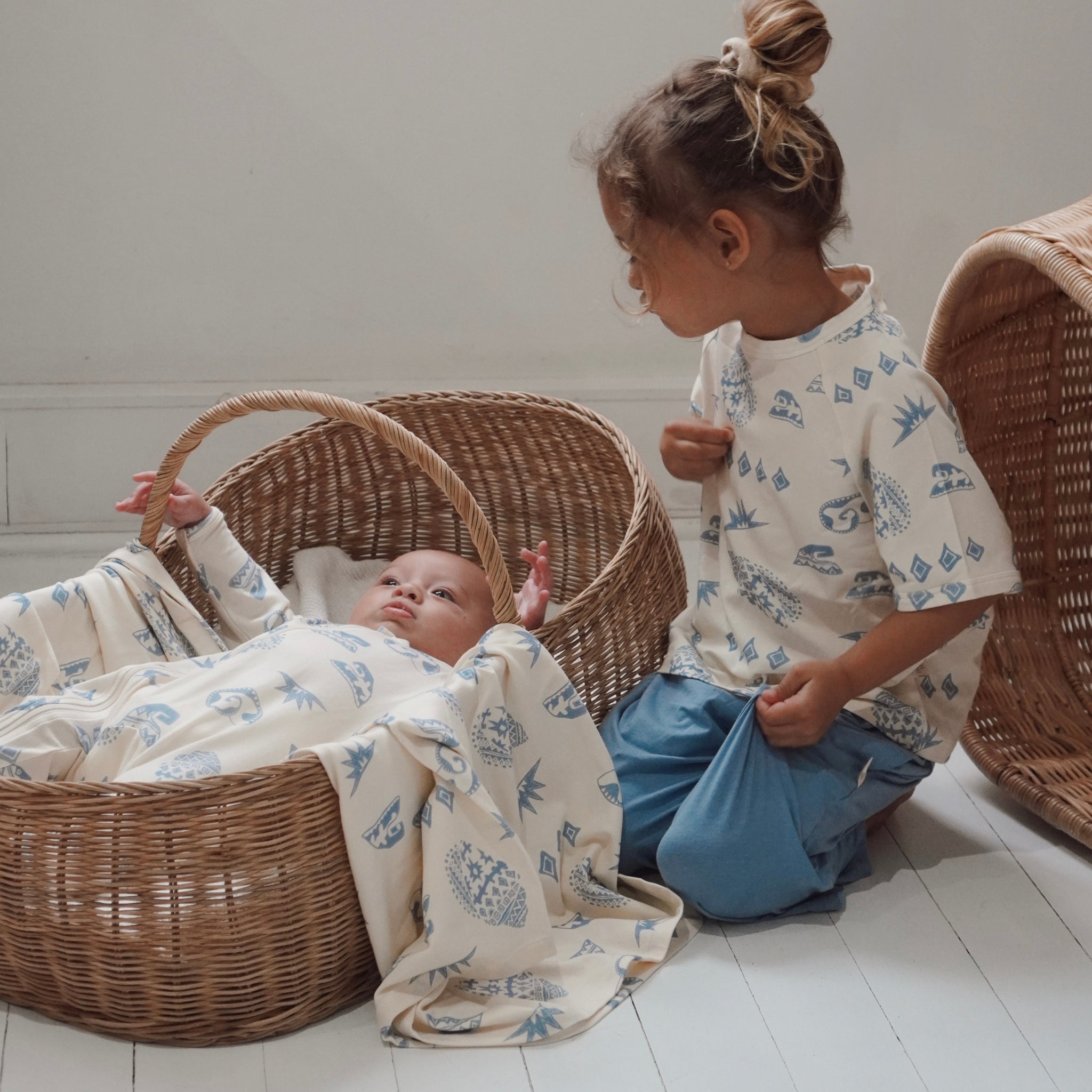 Older child sitting beside baby in a wicker basket, both wearing matching Kailani shell and wave print clothing.