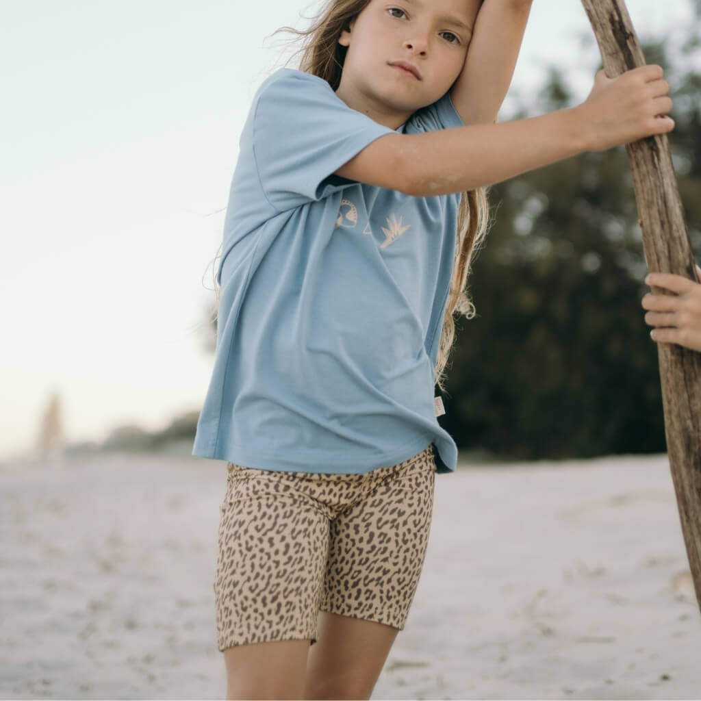 Child wearing a blue shirt and leopard print shorts standing on a beach.