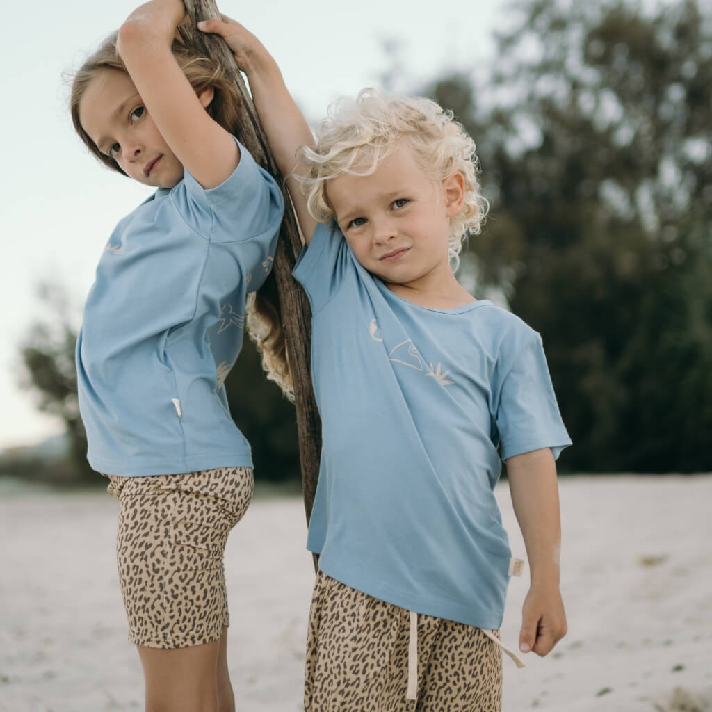 Two children in light blue shirts and leopard print shorts standing on a beach.