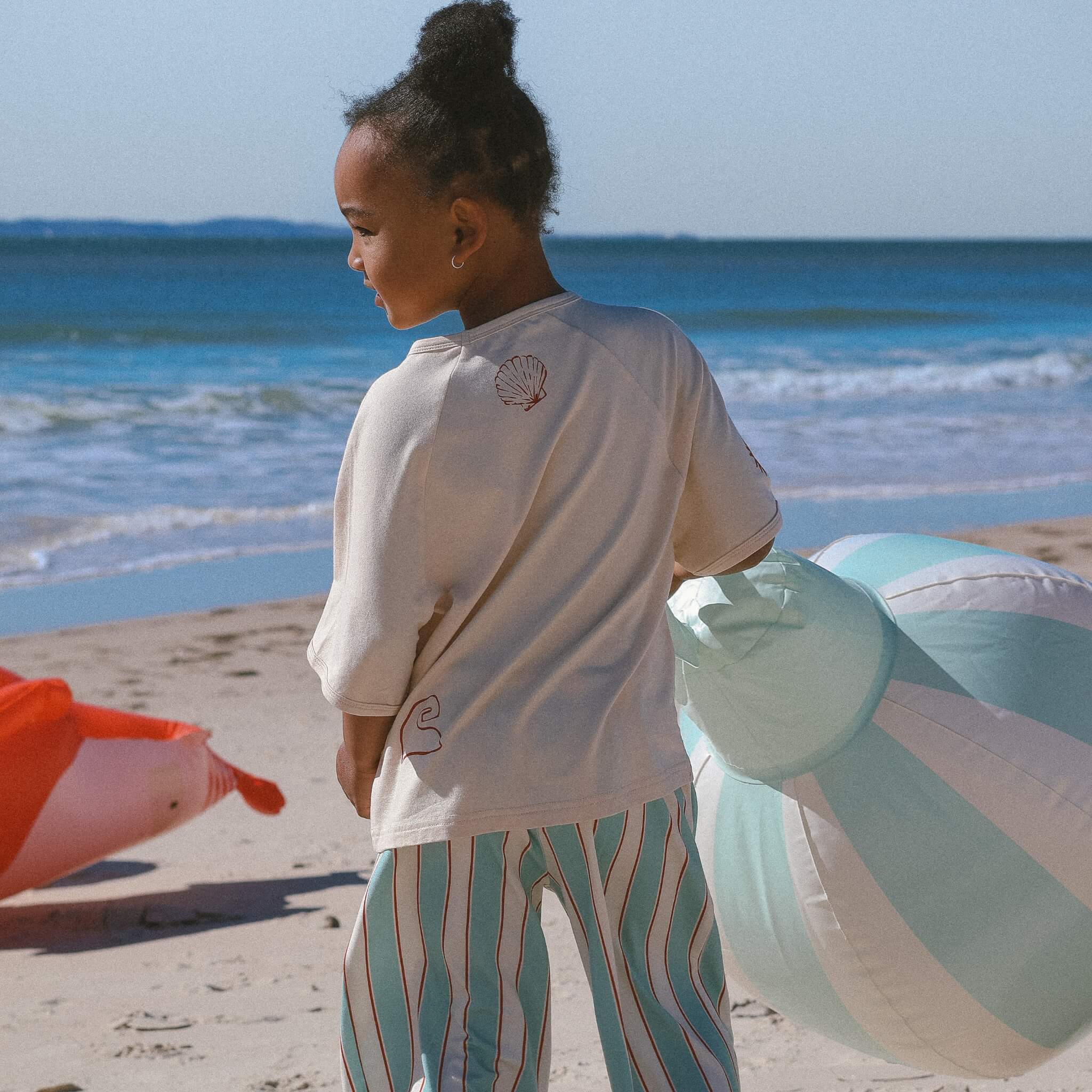 kid in boxy cream tee featuring shell, and wave print paired with mint pinstripe pants on the beach.