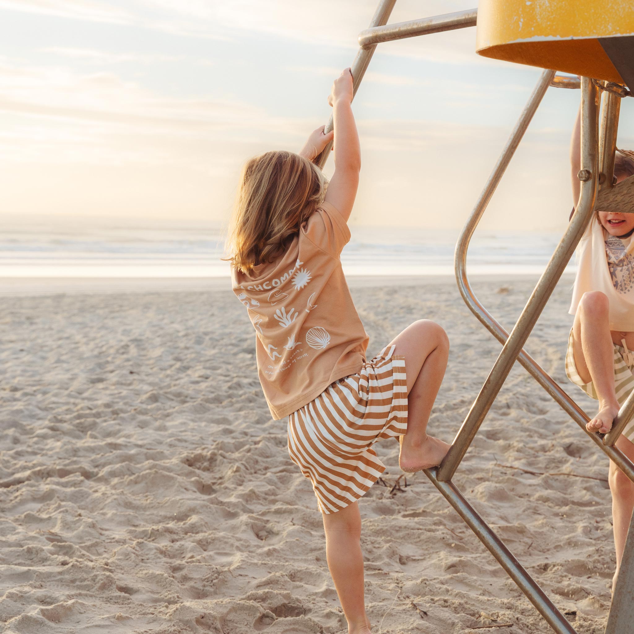 Child climbing a lifeguard tower in a tan Beachcomber print tee by Bam Loves Boo