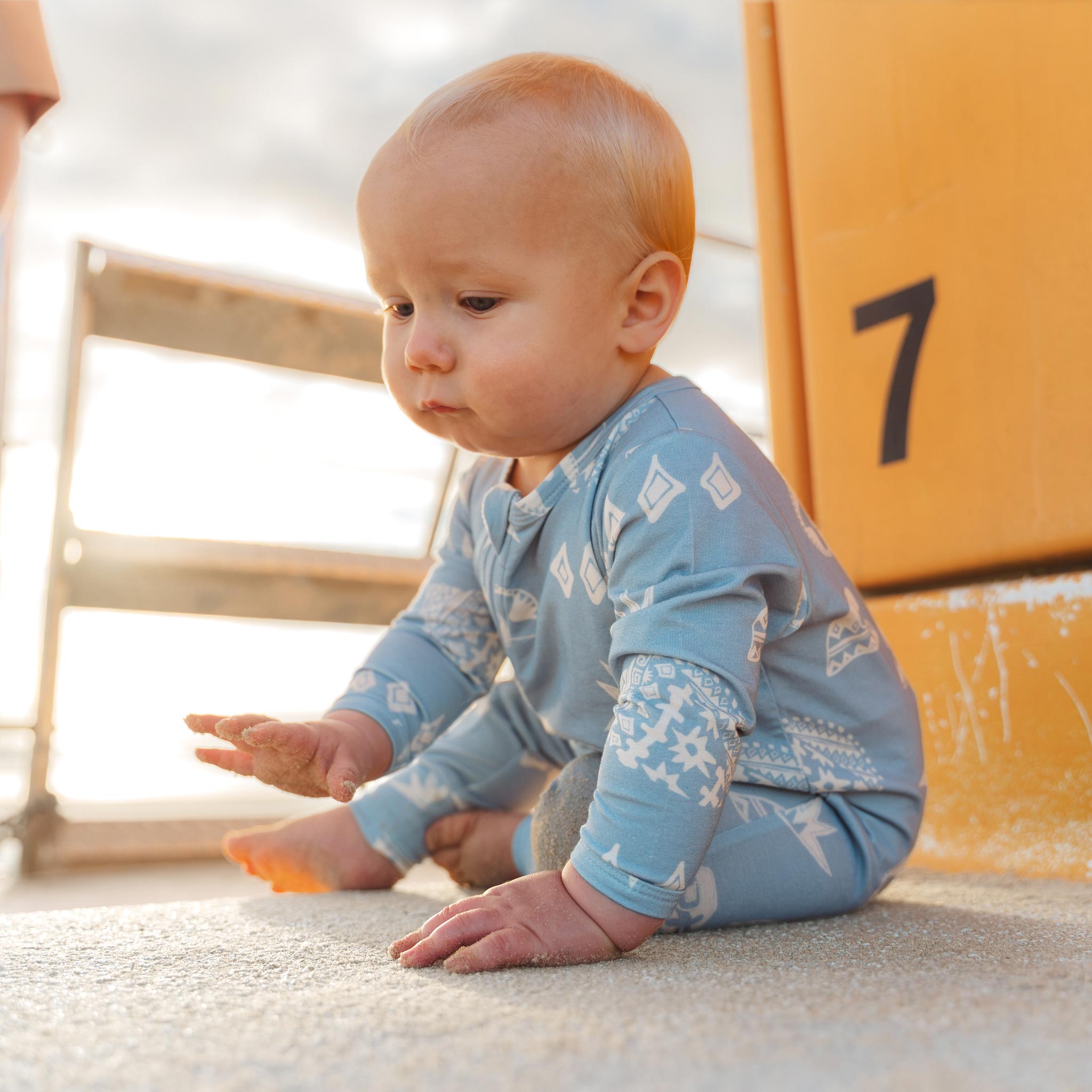 Baby sitting in the sand in a blue Kailani shell print zip jumpsuit by Bam Loves Boo