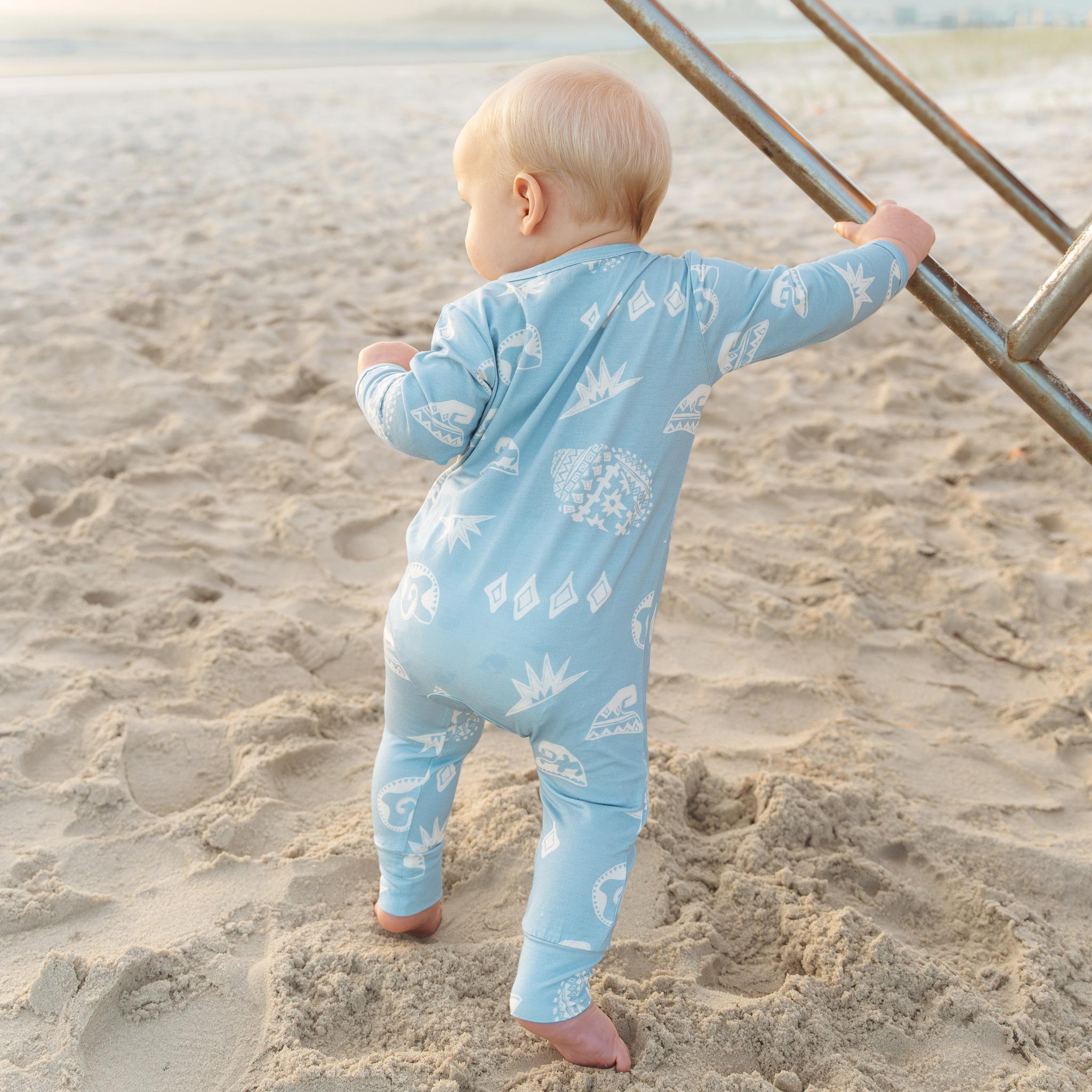 Baby standing on the sand in a blue Kailani shell print zip jumpsuit by Bam Loves Boo