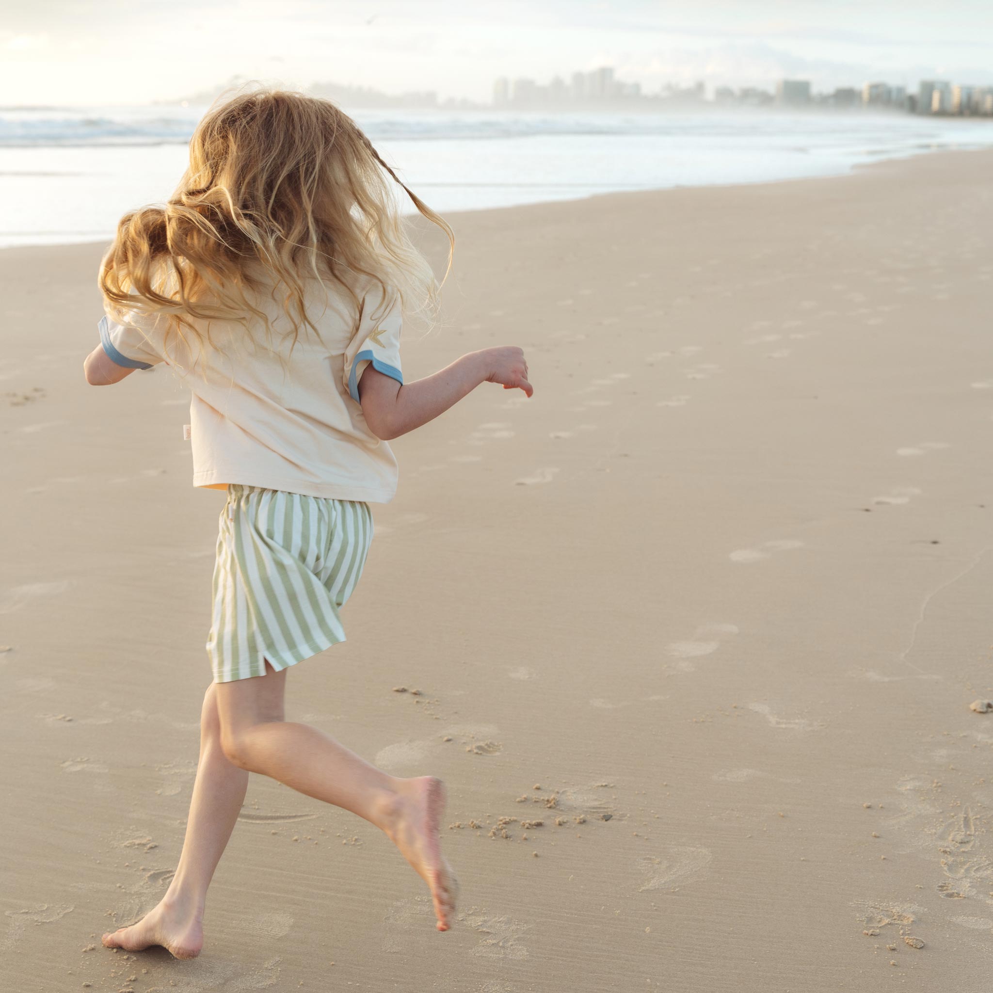 Child running along the beach in a cream Kailani shell print raglan tee by Bam Loves Boo