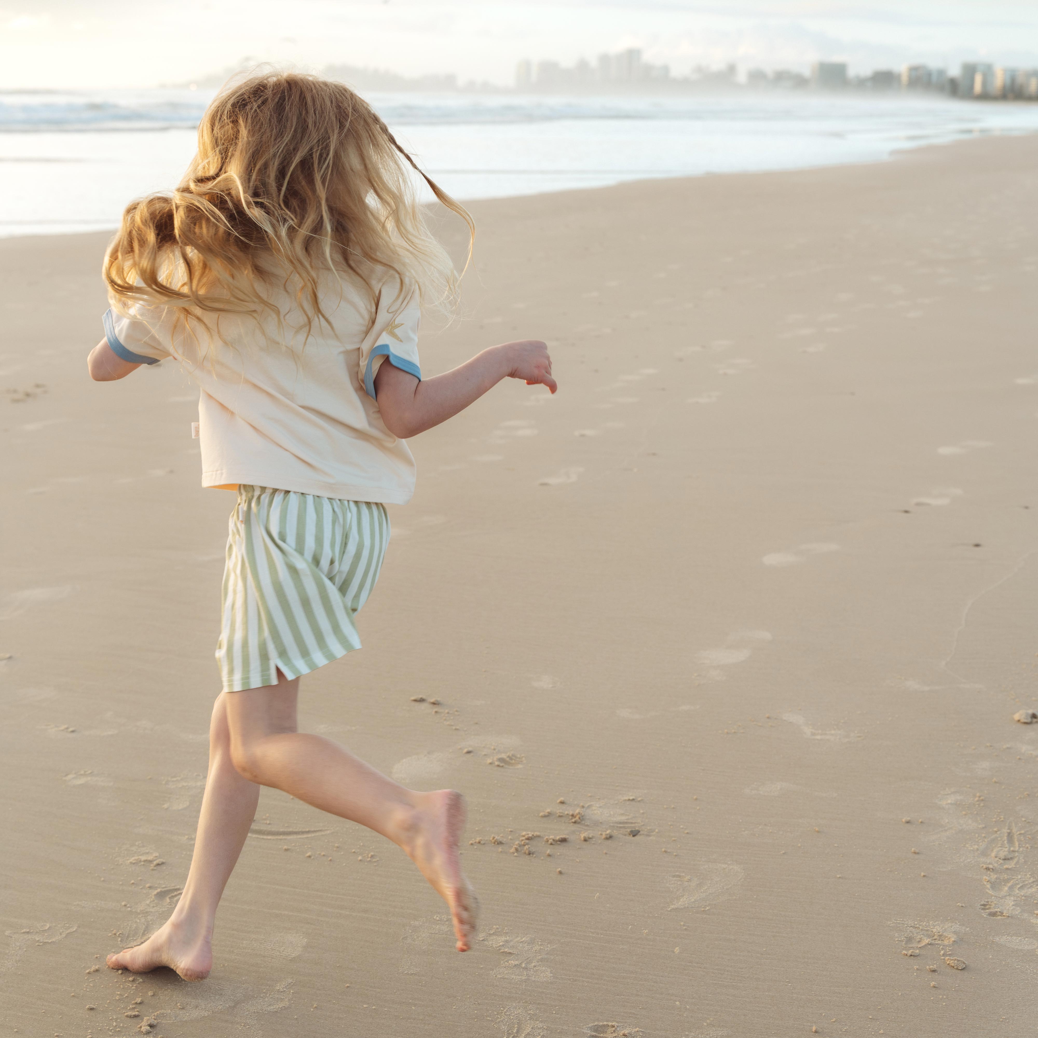 Child running on the sand in sage green and cream striped shorts by Bam Loves Boo