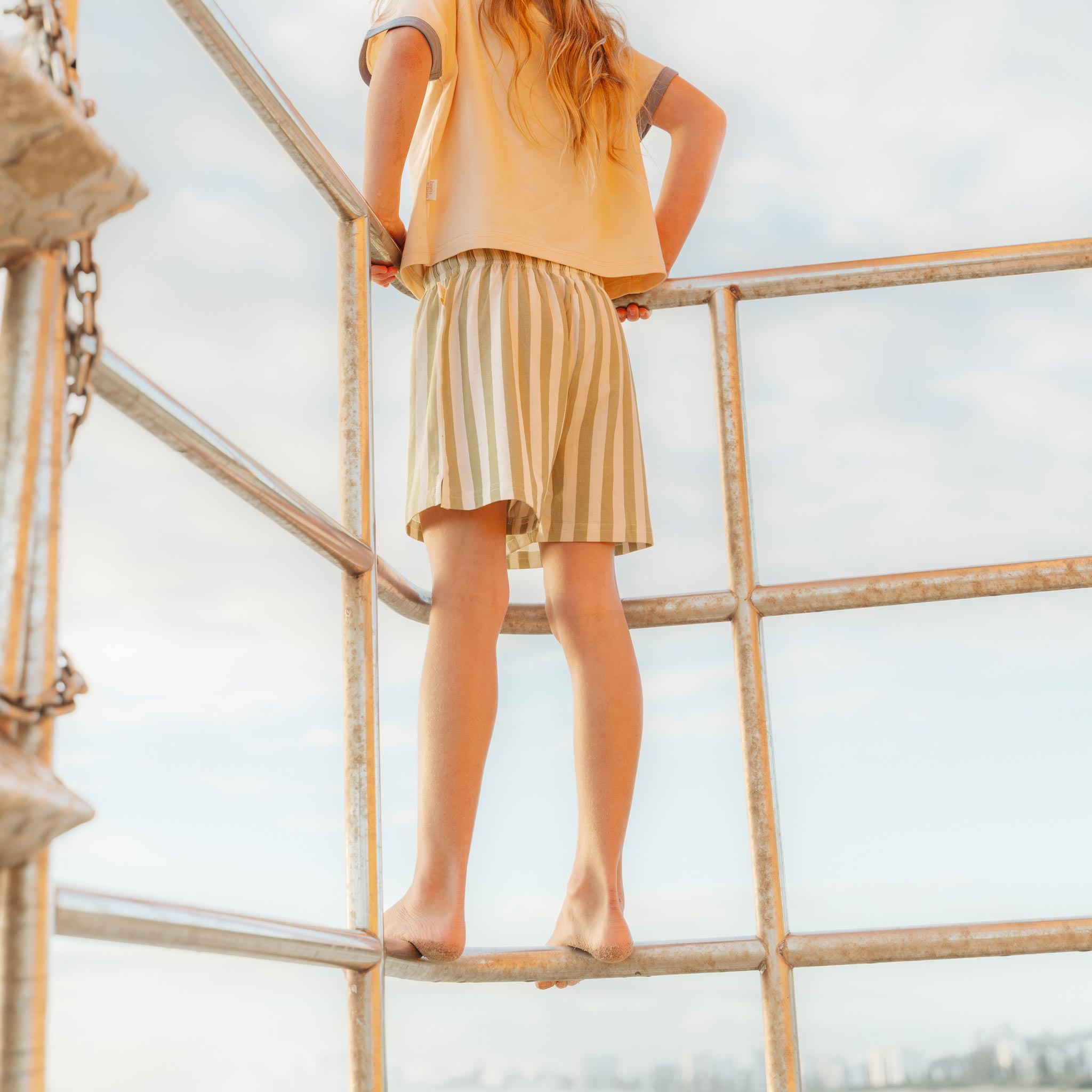 Child climbing a lifeguard tower in sage green and cream striped shorts by Bam Loves Boo